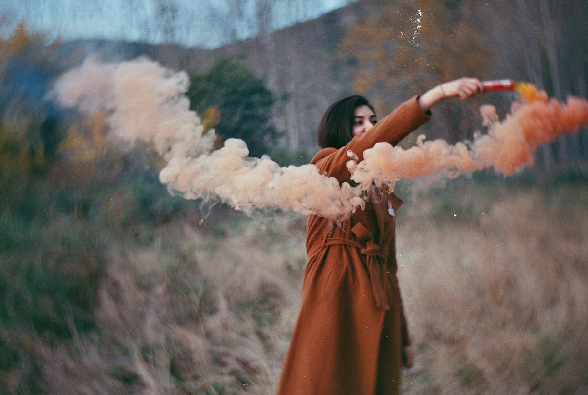 Young woman holds an orange smoke bomb in a misty, autumnal setting outdoors.