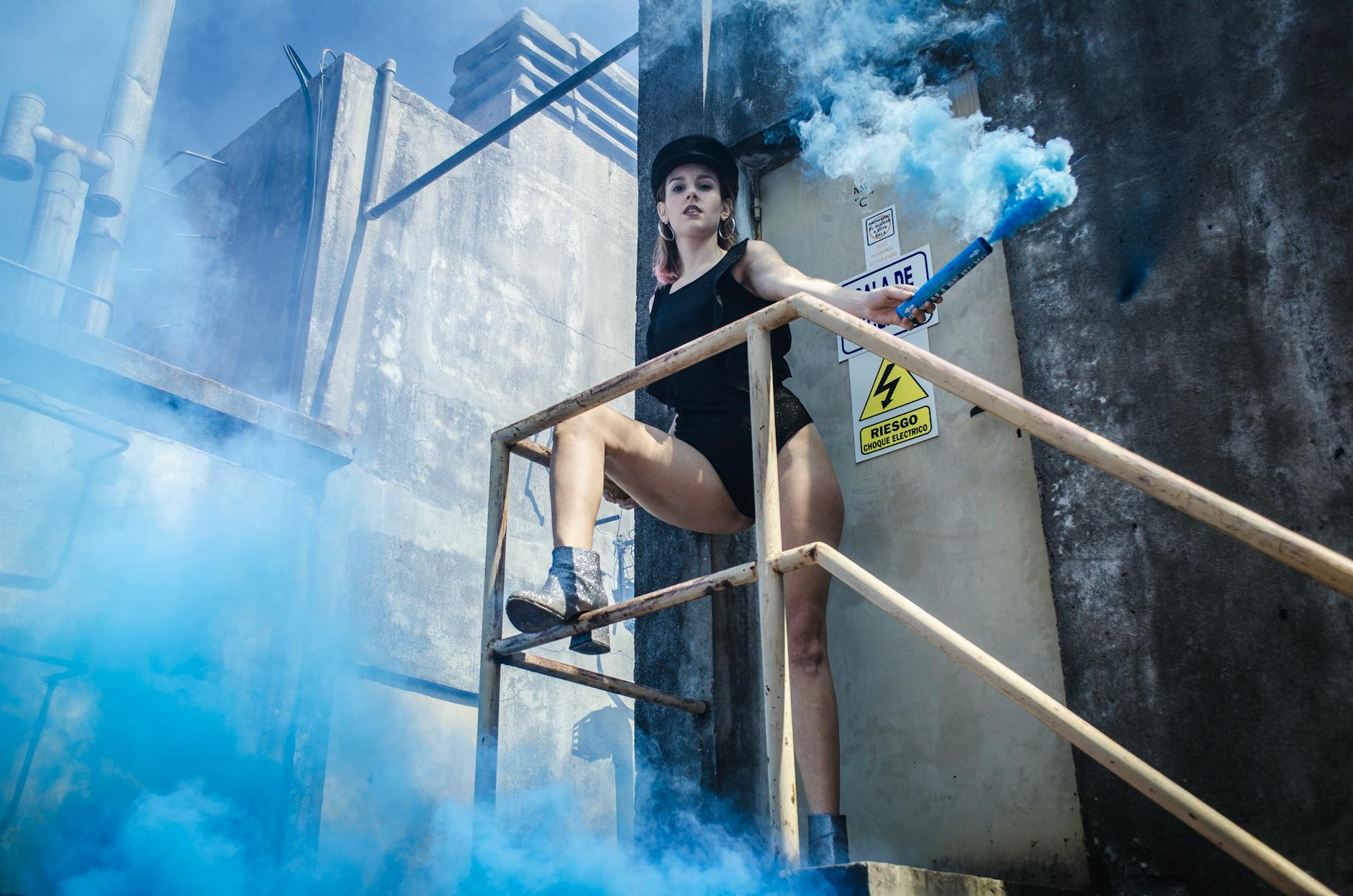 A woman holding a blue smoke bomb on an industrial staircase, creating an edgy fashion scene.