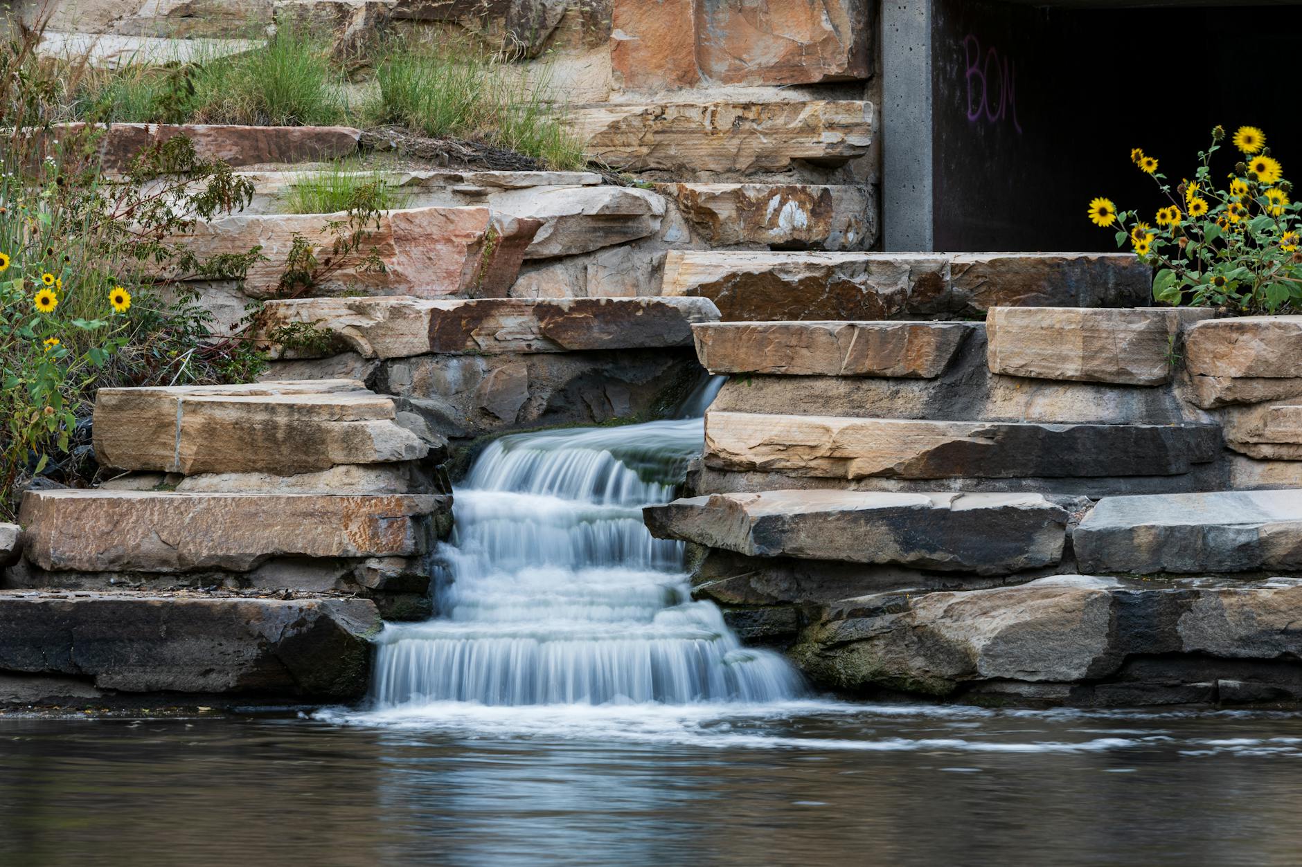 Serene urban waterfall cascading over stones with yellow flowers. Ideal nature scene for relaxation themes.