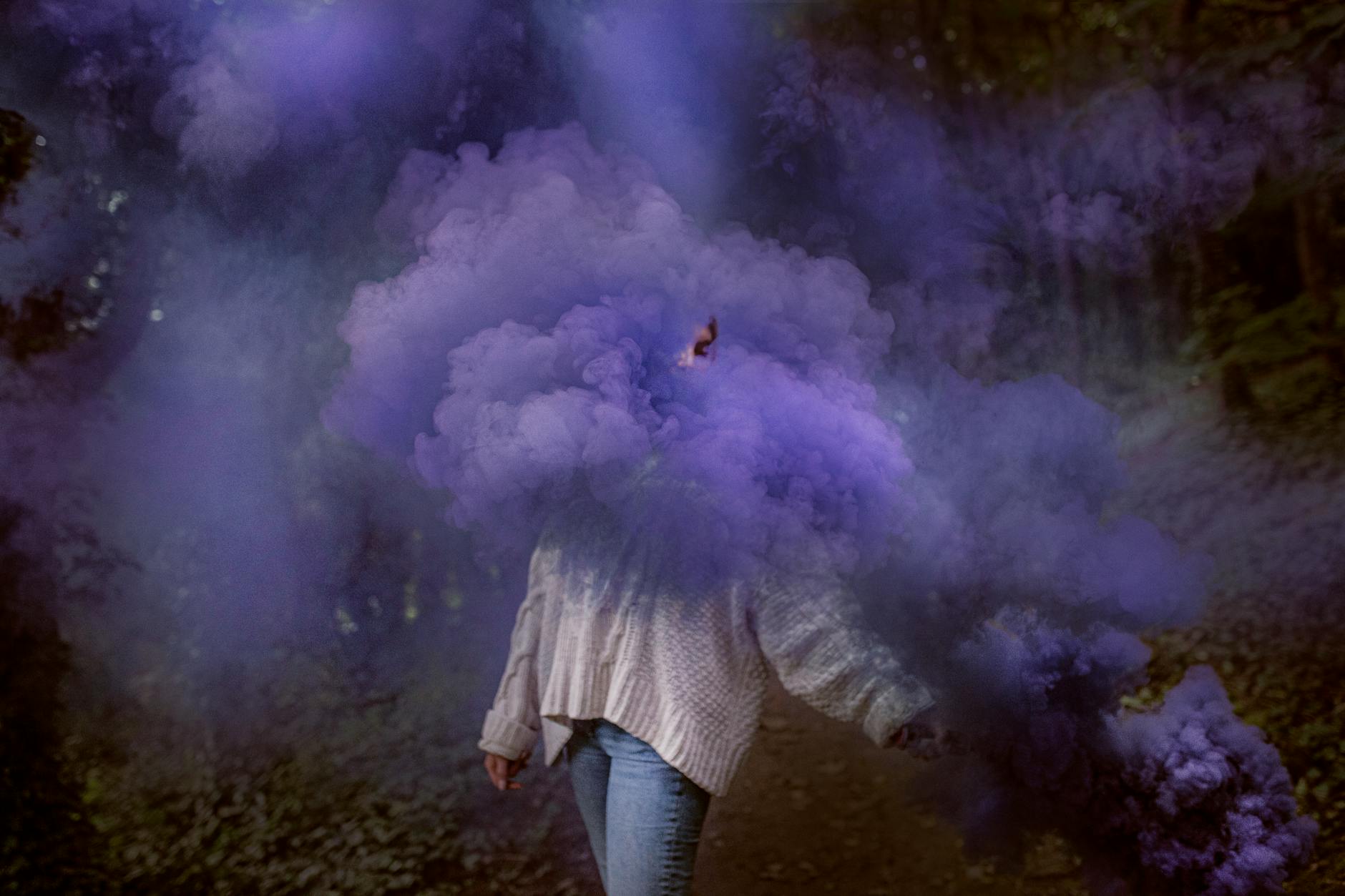 A woman enveloped in vibrant purple smoke in a serene forest setting.