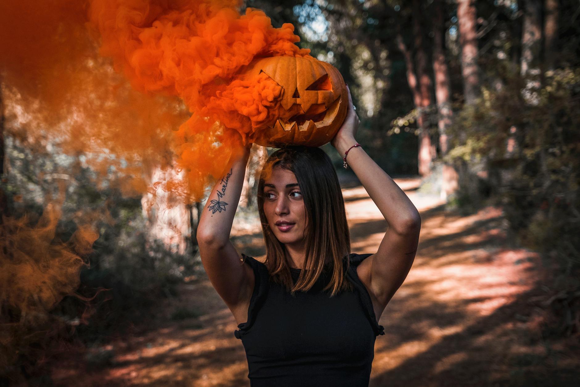 A woman in a forest holding a carved pumpkin with orange smoke, evoking a spooky Halloween mood.