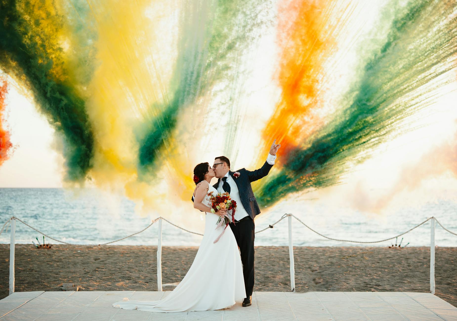 A couple shares a kiss on the beach with vibrant colored smoke creating a stunning backdrop.