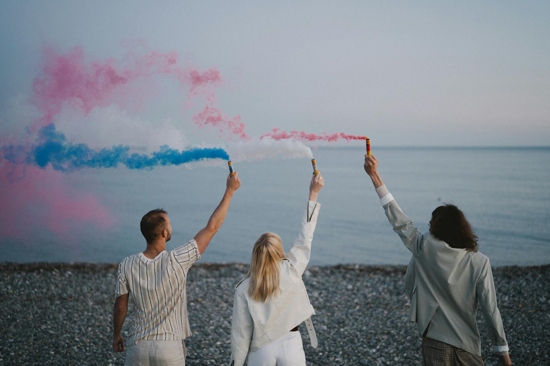 A group of friends with smoke bombs on a stony beach, creating a colorful atmosphere.