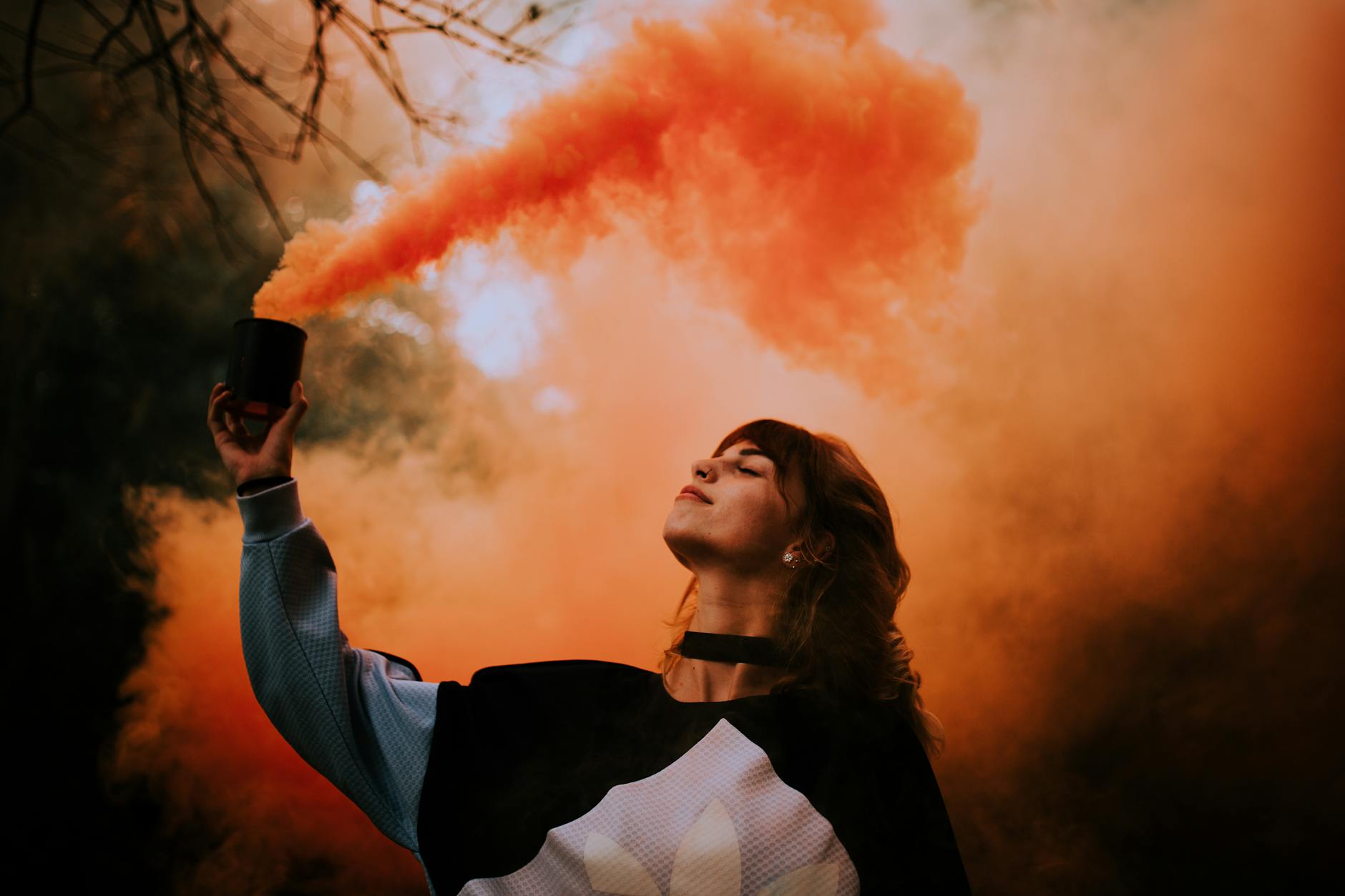 A woman with closed eyes holds an orange smoke bomb outdoors, creating a vibrant and dramatic atmosphere.