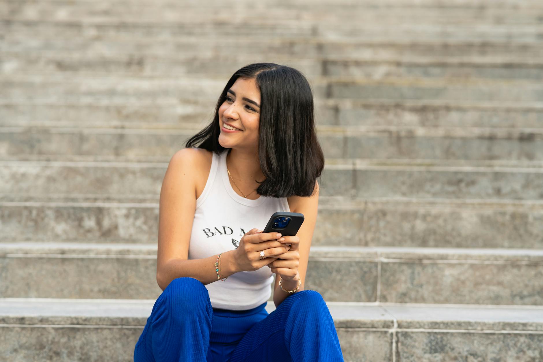 Happy young woman in casual attire enjoying a sunny day outdoors, sitting with a smartphone in Mexico City.