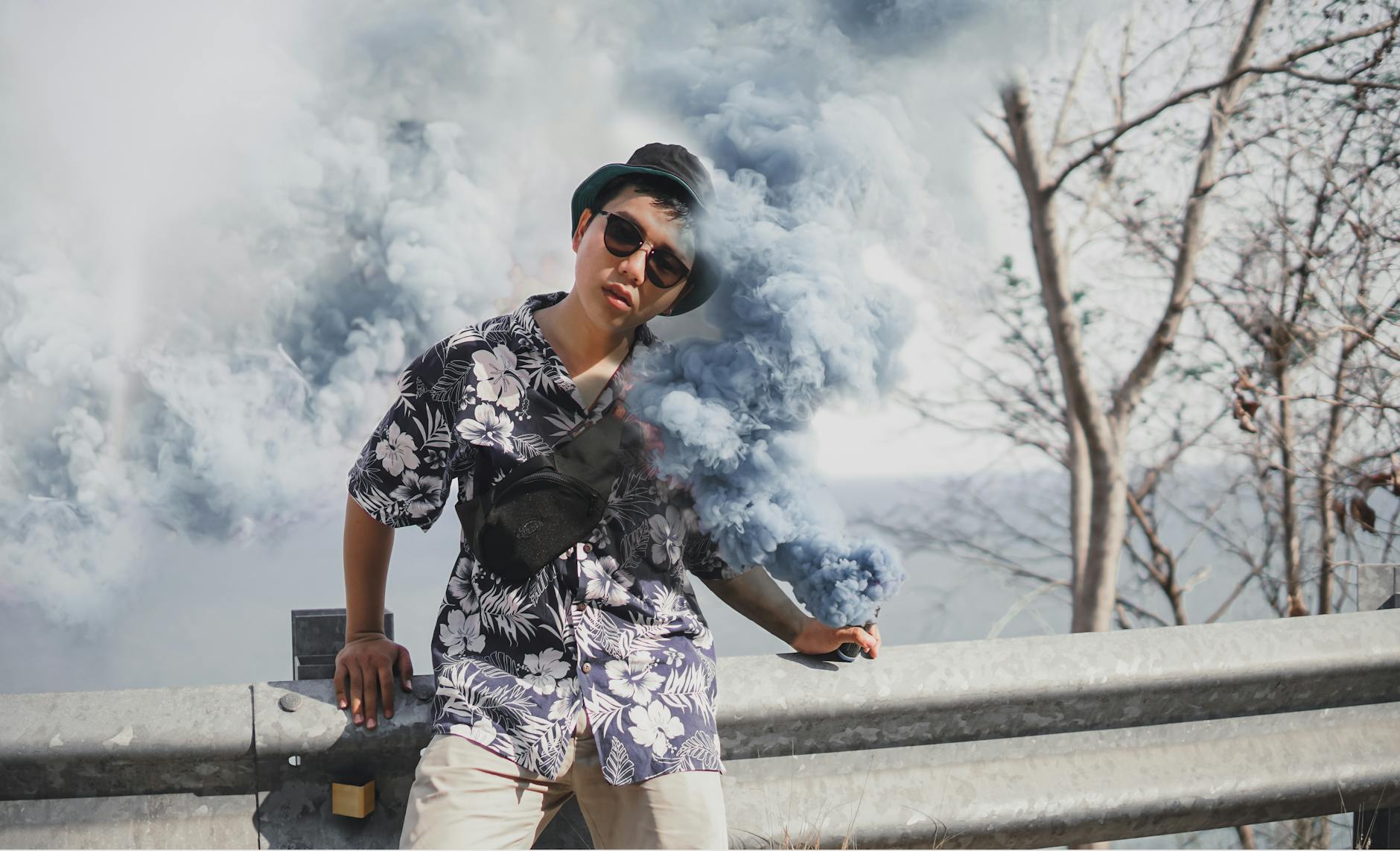 From below of young ethnic male in stylish wear and sunglasses standing near fence with colored smoke spray creating vapor effect in air