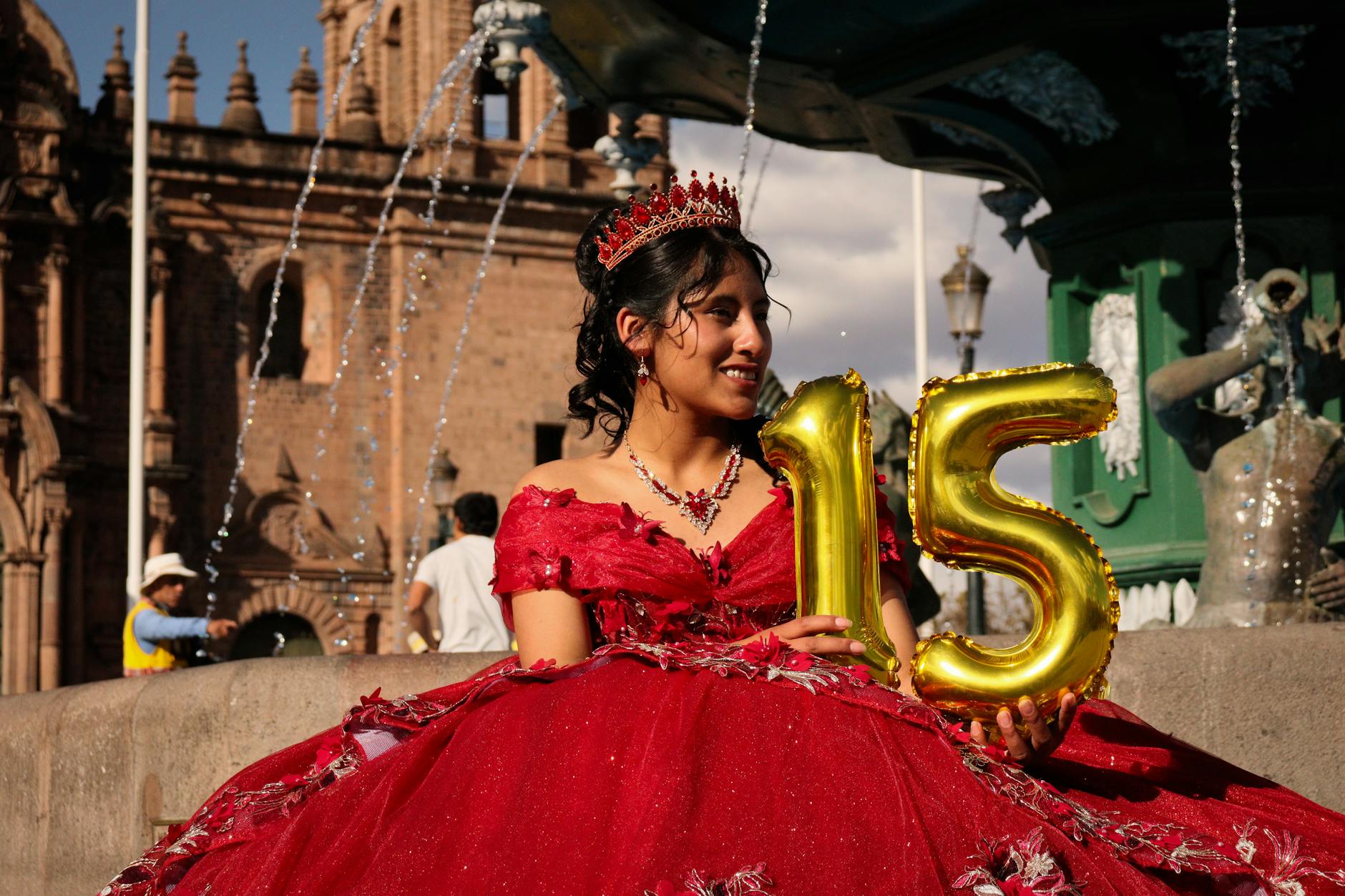 A young woman celebrates her 15th birthday in a vibrant red gown by a historic fountain.