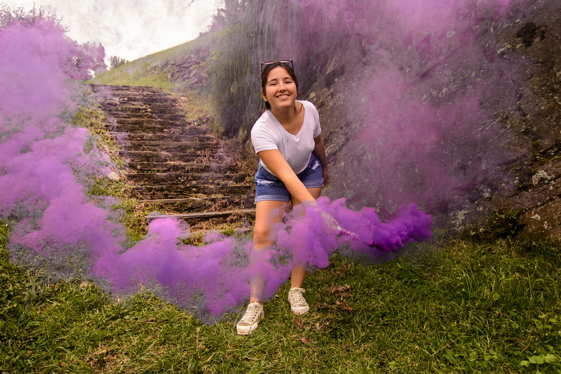 A cheerful young woman playfully holding a purple smoke bomb on stone steps in an outdoor setting.