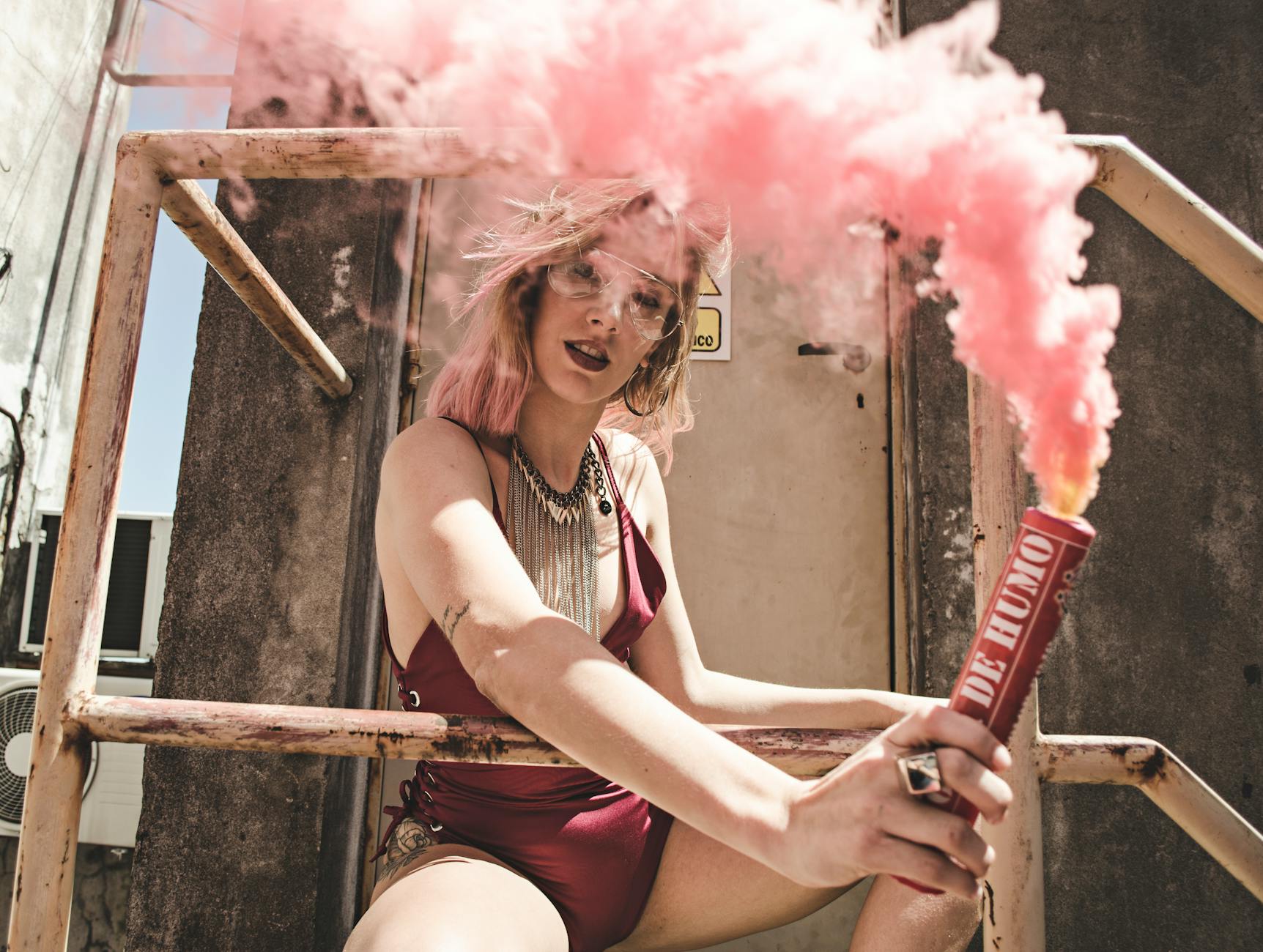 Stylish young woman in red, holding pink smoke grenade outdoors on staircase.