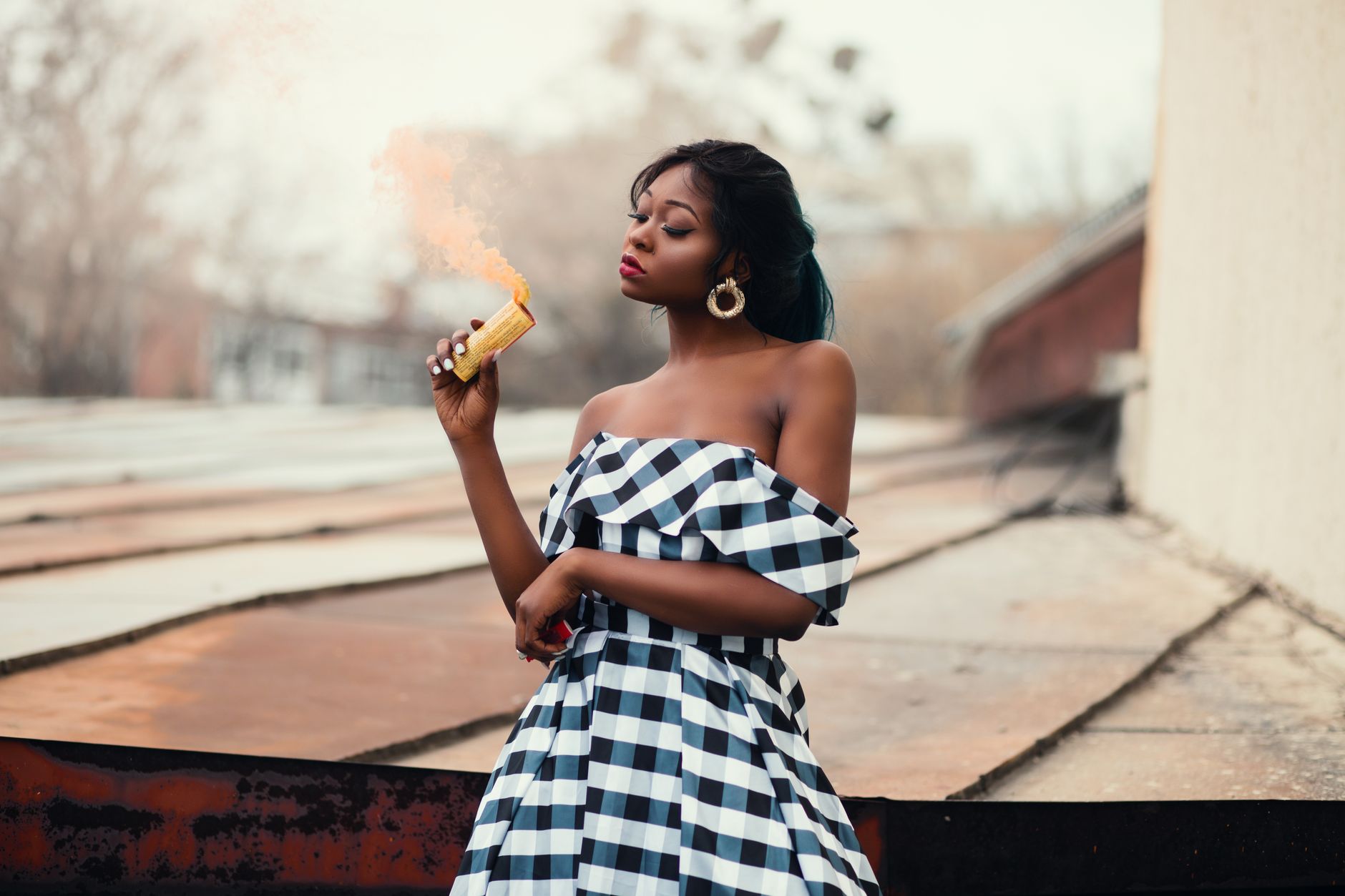 Fashionable woman stands on rooftop holding smoke flare in a stylish checkered dress.