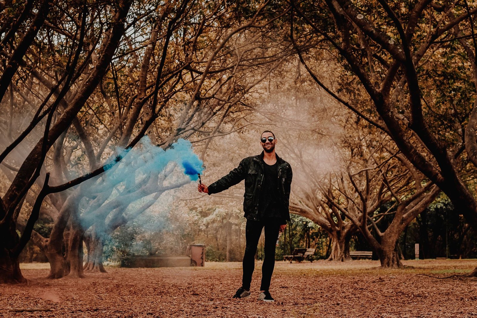 A fashionable man poses with a blue smoke bomb amidst a forest setting.