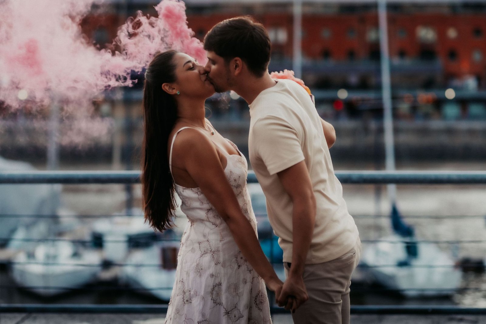A young couple shares a romantic kiss by the waterfront in Buenos Aires, enveloped in pink smoke.