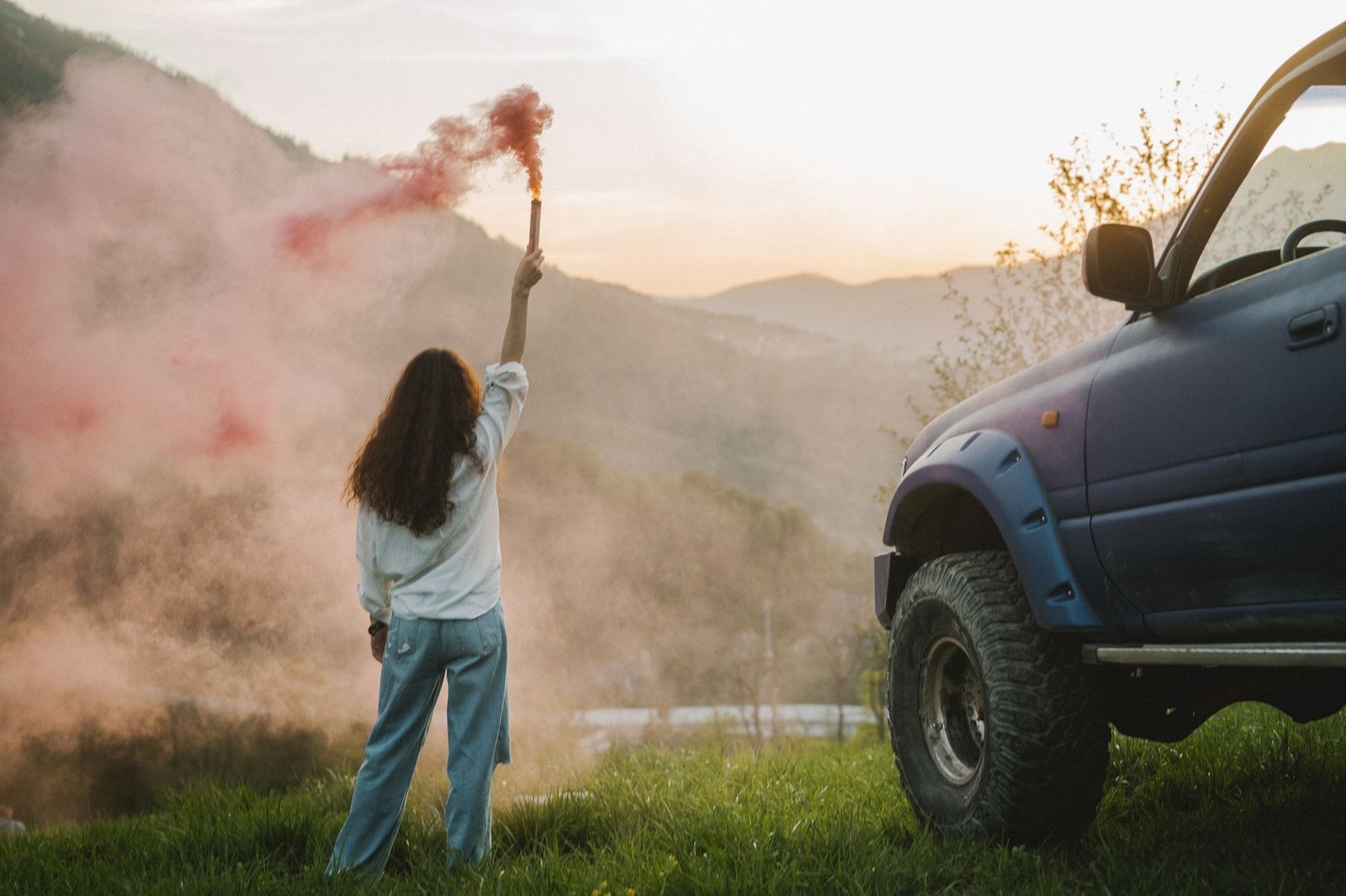 Woman with smoke flare and SUV in scenic mountain landscape at sunset.