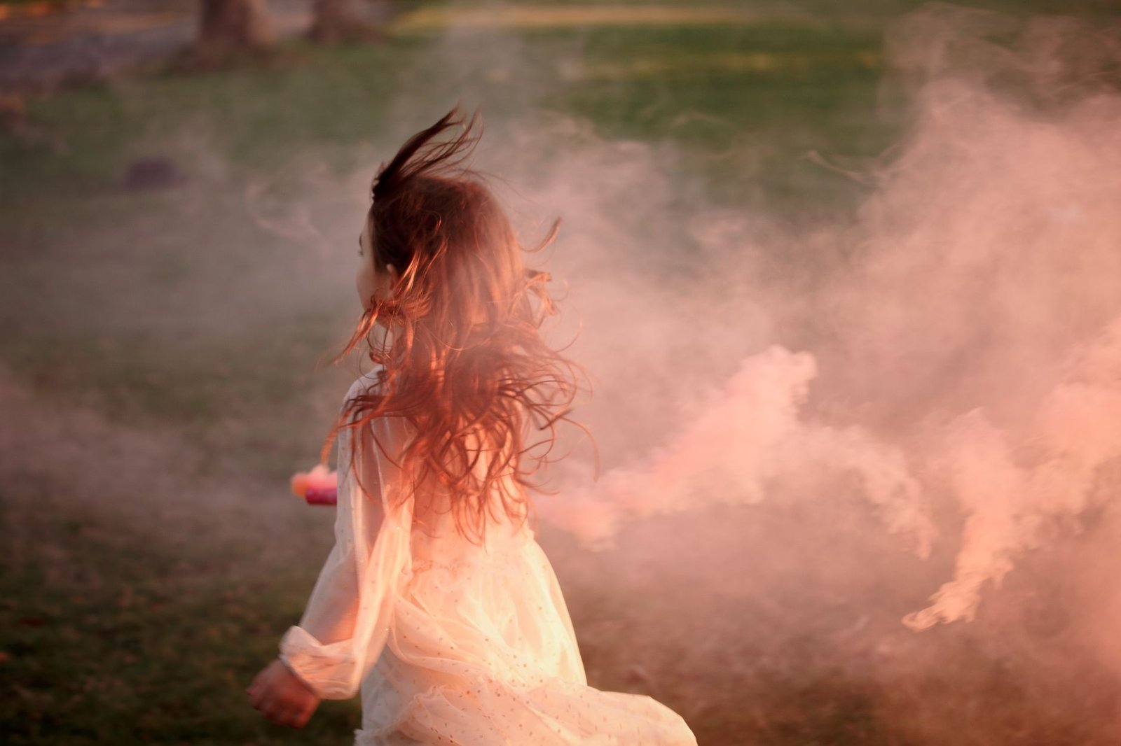 A young girl in a white dress playing in a misty field at sunset, surrounded by smoke.
