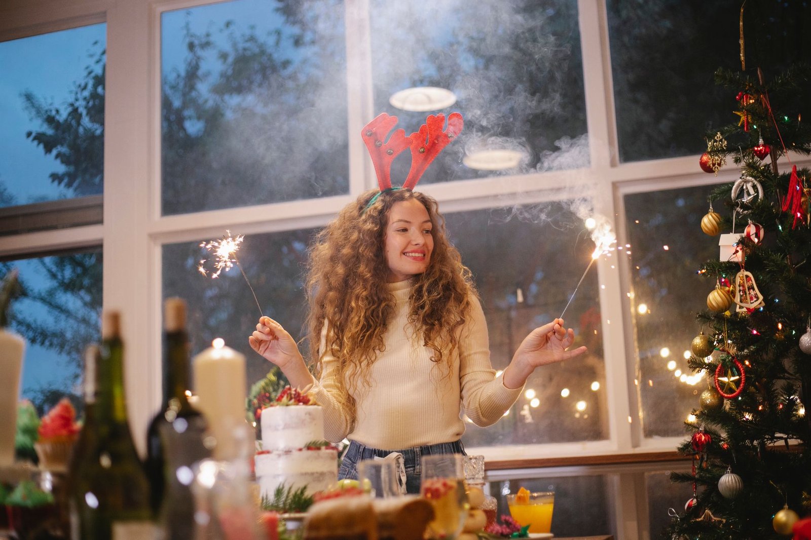 Low angle of smiling female in deer headband with burning sparklers standing near festive table and decorated fir tree and celebrating Christmas