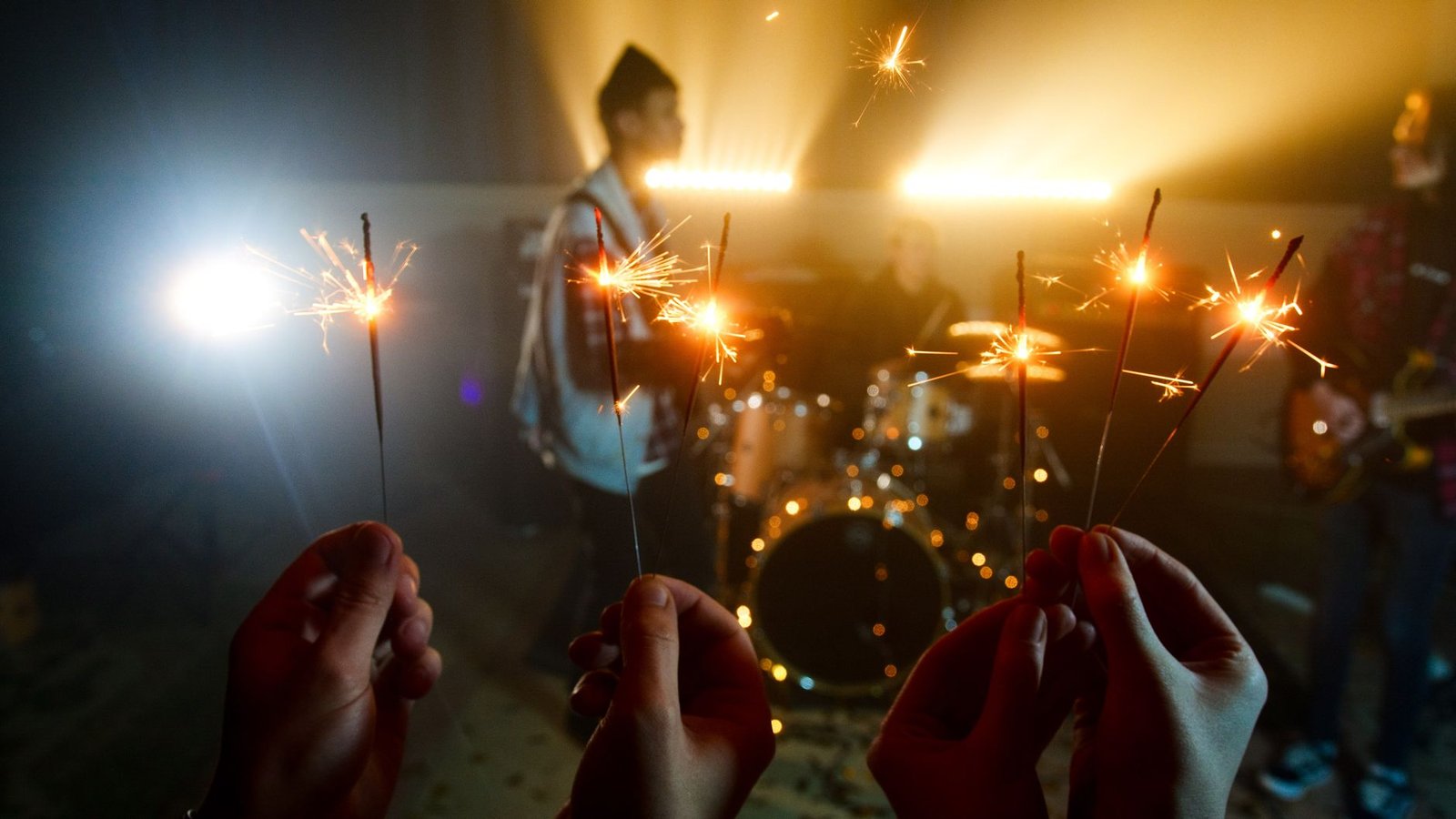 Hands holding sparklers in front of a band performing indoors, creating a festive atmosphere.