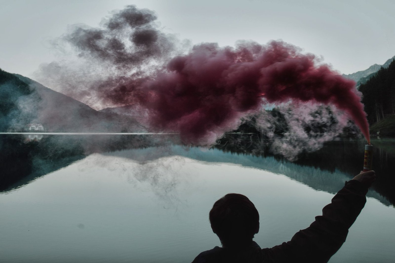 Silhouette holding smoke bomb by a tranquil lakeside with mountain reflection.