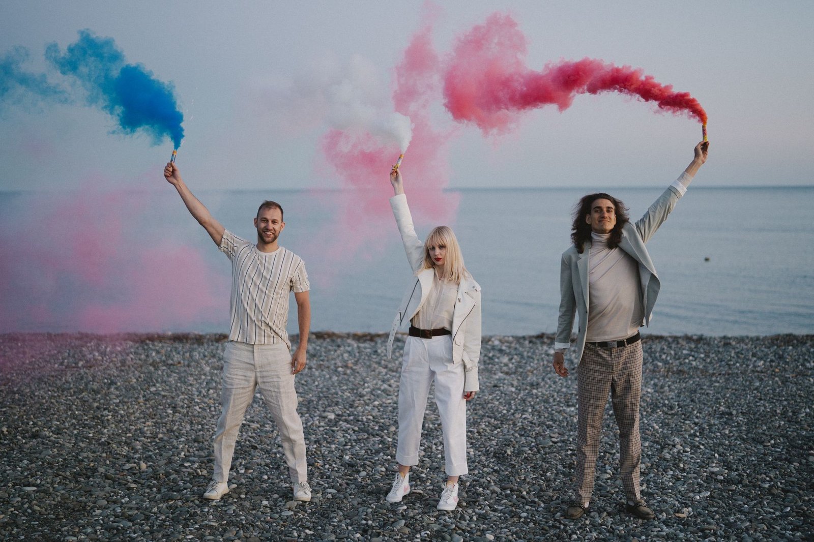 Three adults holding vibrant smoke bombs on a pebble beach by the sea.