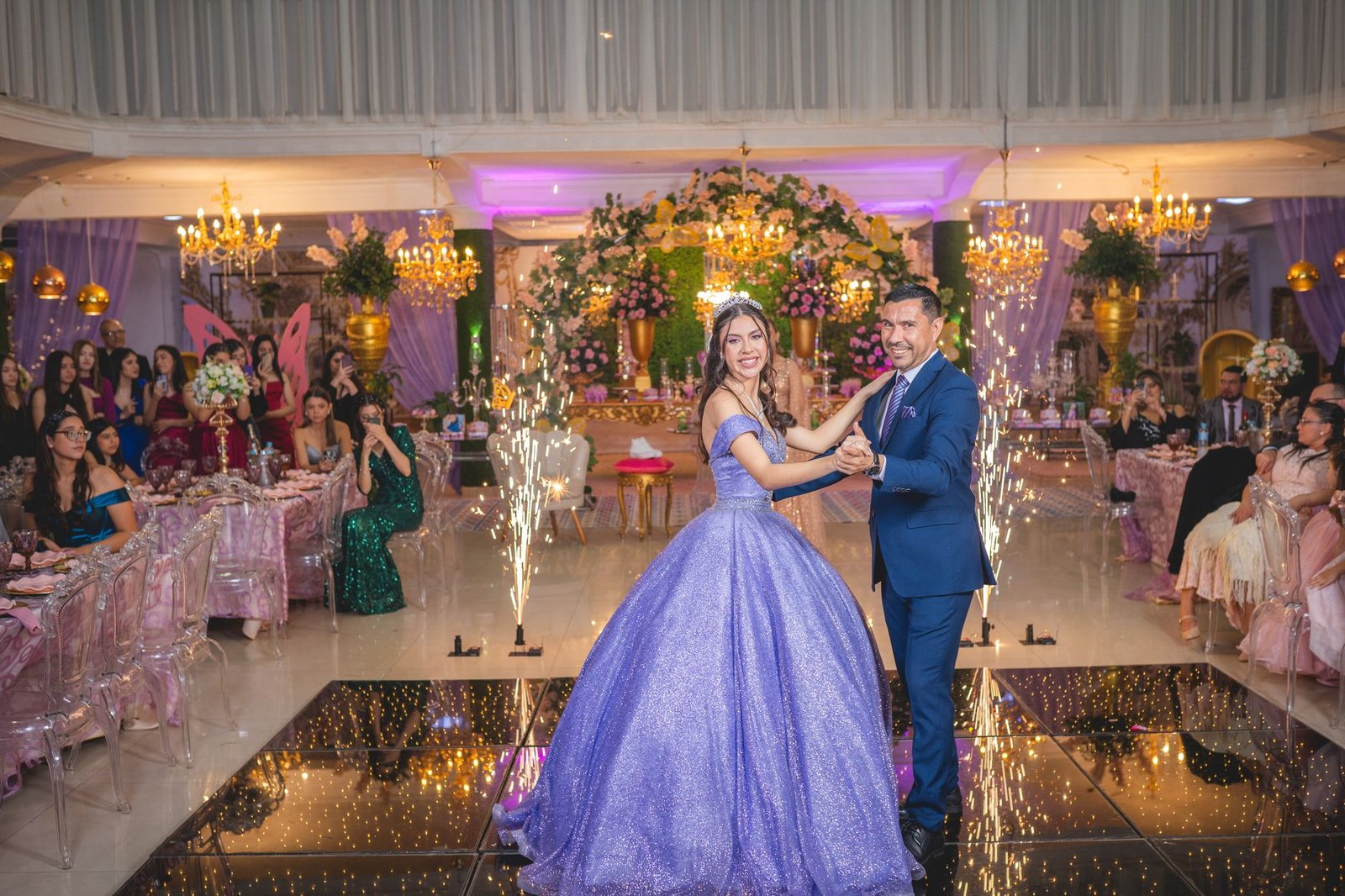 A couple elegantly dances at a lavish quinceañera celebration amidst joyful guests and sparkling decor.