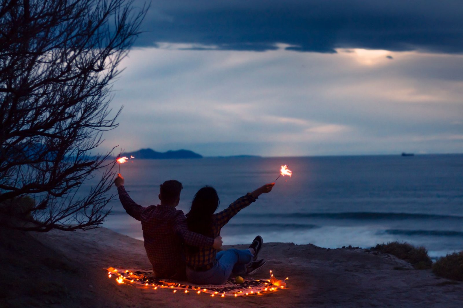 Silhouette of a couple with sparklers sitting by the ocean at sunset, creating a romantic mood.
